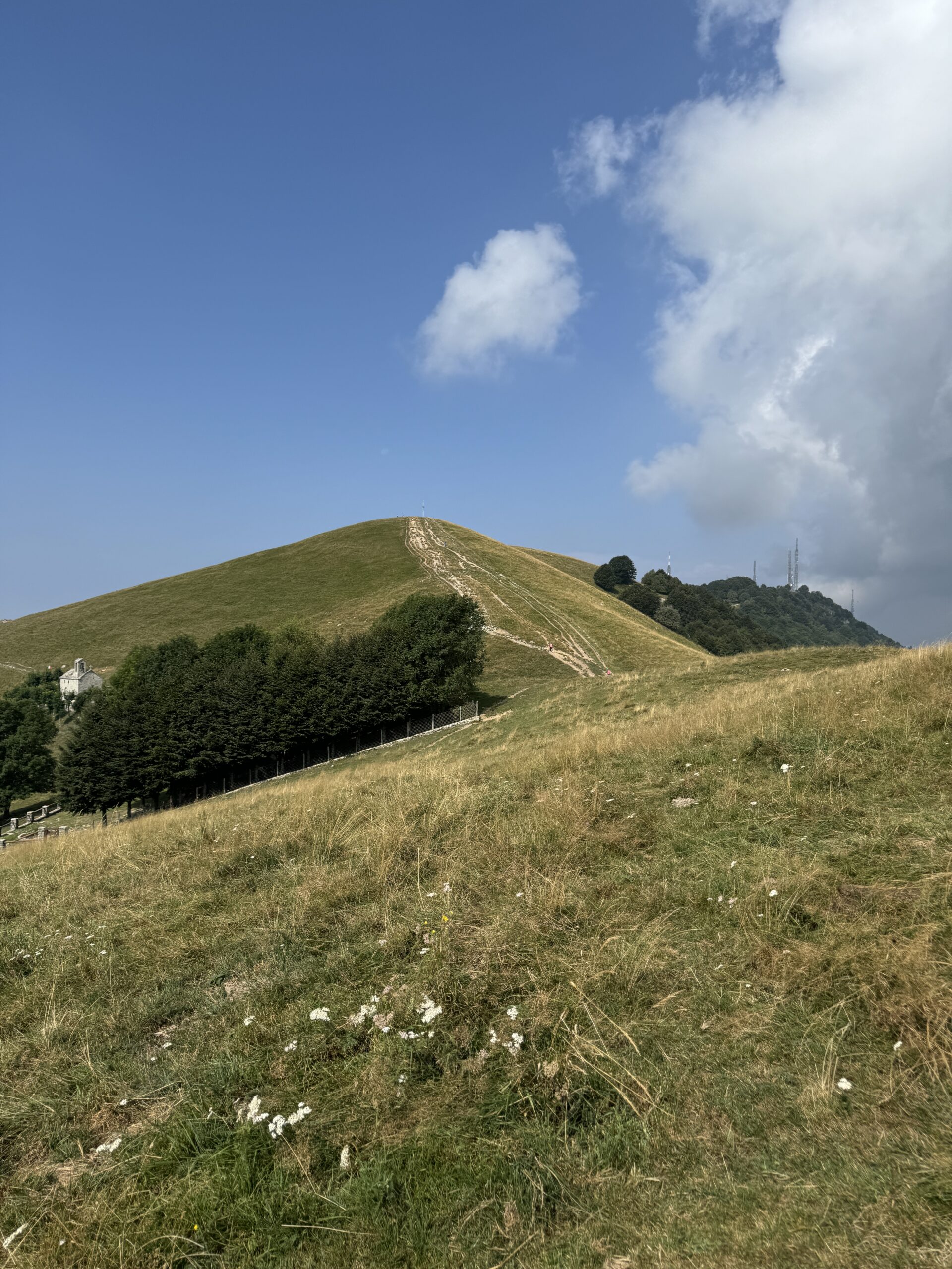 Sentiero erboso che sale al Monte Linzone da Valcava, con cielo azzurro e nuvole sullo sfondo.